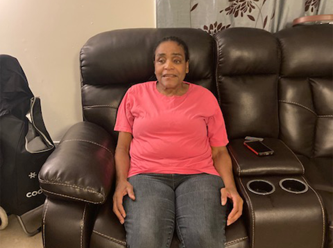 Mary Conner sitting on her couch, wearing a bright pink shirt and jeans.