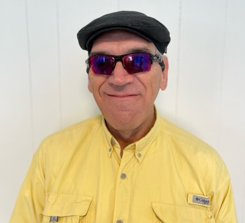 Randall Tibbett smiling for the camera, wearing sunglasses, a cap, and a bright yellow shirt.