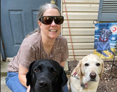 Judy Davis, wearing a light brown shirt, jeans, and sunglasses, kneeling on the walkway in front of her house with her two dogs. Orville, a black Lab, is in front, and Prince, a yellow lab, is to the right