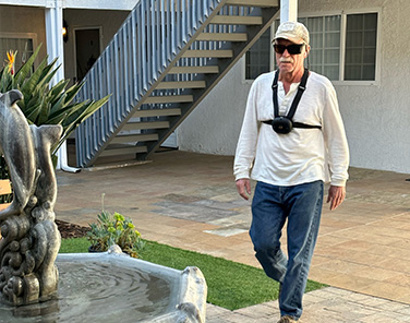 Ron Peterson, walking across a courtyard, toward a fountain. Ron is wearing an innovative navigation device called Ara, which scans the environment and provides feedback about obstacles by creating vibrations in the straps. The device is black, about the size of a fist, and the straps are fastened around the chest and over the shoulders.
